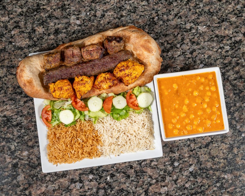 A platter of grilled meats, rice, salad, and a side of chickpea stew on a granite surface.