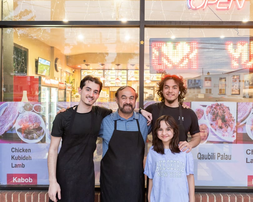 Four smiling people, three men and one child, stand in front of a restaurant window with food posters.